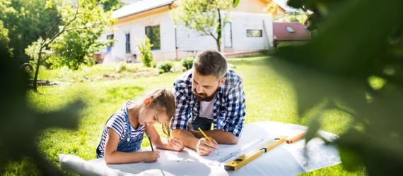 Bausparrechner: Vater zeichnet mit Tochter auf Wiese vor Haus an einem Bauplan.