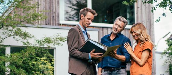 Paar und Berater stehen vorm Haus und schauen sich Unterlagen an