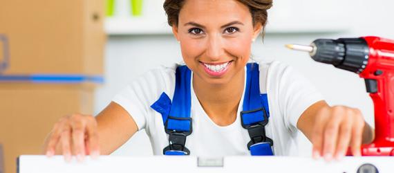 Junge Frau mit weißem T-Shirt und Blaumann hält Wasserwaage. Daneben steht eine Bohrmaschine.