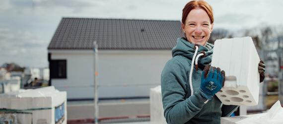 Junge Frau auf Hausbaustelle hält einen Kalksandstein in den Händen