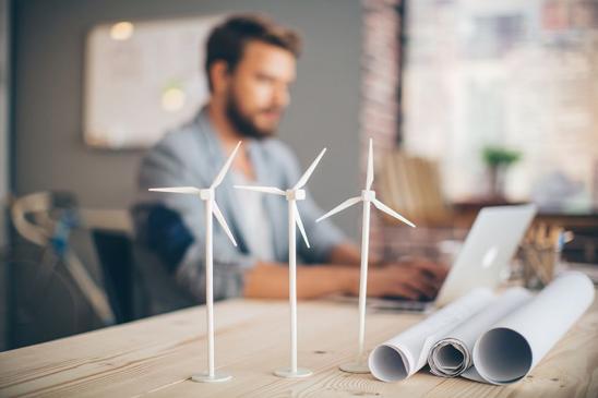 Architekt sitzt am Schreibtisch bei der Planungsarbeit. Im Fokus: zusammengerollte Baupläne und Windradmodelle